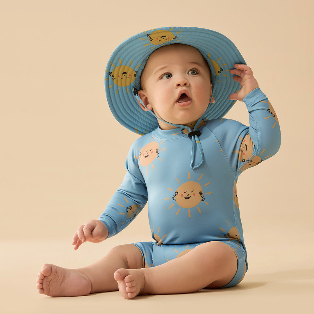 Baby wearing a blue sun protective outfit with sun designs and a matching hat on a beige background